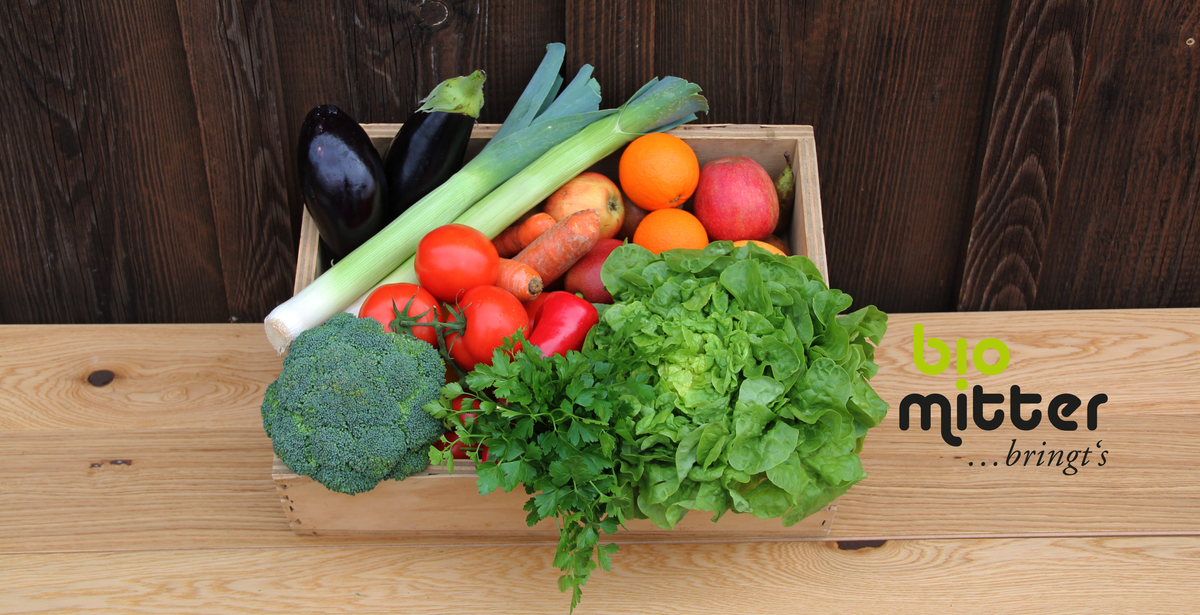 KI generiert: Das Bild zeigt eine Kiste mit frischem Gemüse und Obst, darunter Brokkoli, Tomaten, Salat und Äpfel, vor einem Holzhintergrund. Daneben ist das Logo von "bio mitter" abgebildet.