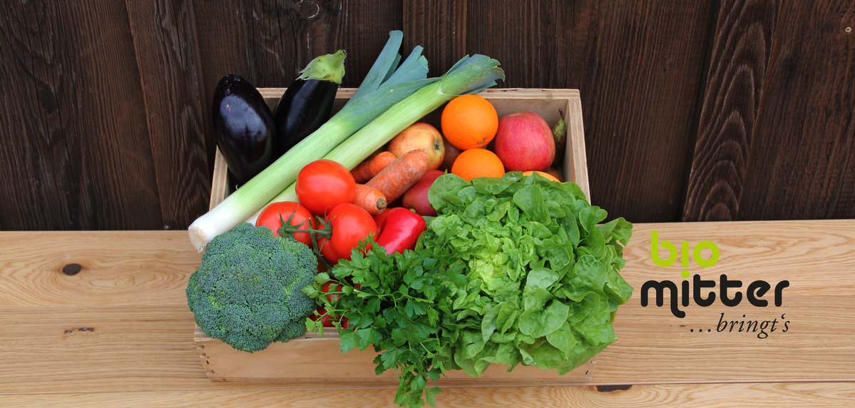 KI generiert: Das Bild zeigt eine Kiste mit frischem Gemüse und Obst, darunter Brokkoli, Tomaten, Salat und Äpfel, vor einem Holzhintergrund. Daneben ist das Logo von "bio mitter" abgebildet.