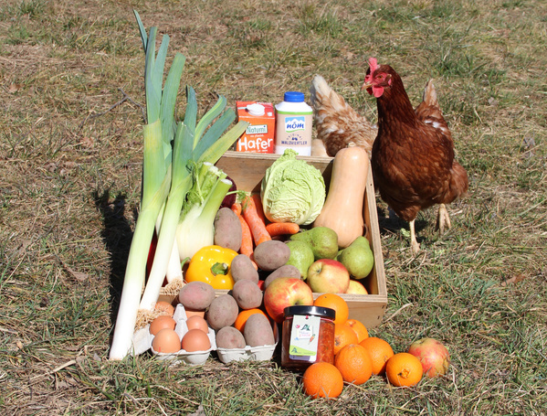 KI generiert: Eine Holzkiste mit Obst, Gemüse, Eiern, Milch und Haferdrink auf Gras, daneben ein Huhn.