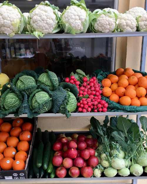 KI generiert: Verschiedene Gemüsesorten und Früchte auf Regalen; Blumenkohl, Kohl, Radieschen, Orangen, Gurken, Äpfel.
